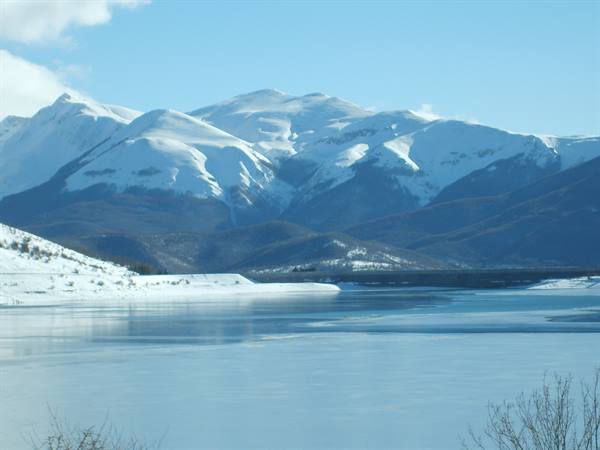 Frozen lake before snowy mountains, snow-covered shore and blue sky