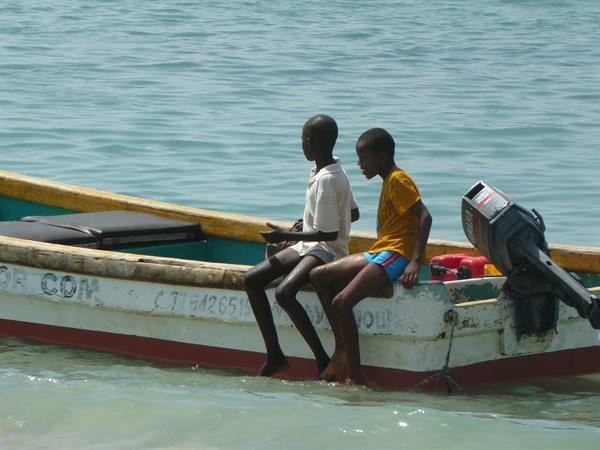 Two boys sitting on the edge of a wooden boat with outboard motor, feet in turquoise water