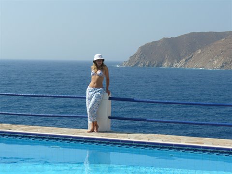 Woman in hat leaning on white pillar by the pool, sea and cliffs behind