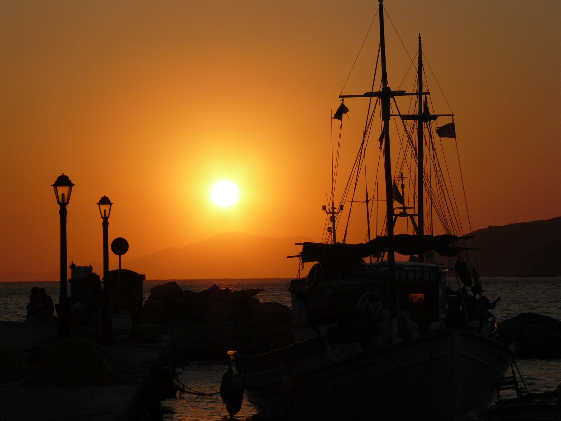 Veleiro in silhouette al molo con lampioni, sole arancio sul mare