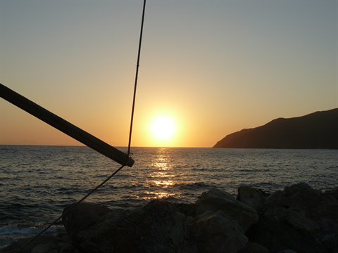 Low sun over the sea behind rocks and a dark mountain, boat mast spar in the foreground