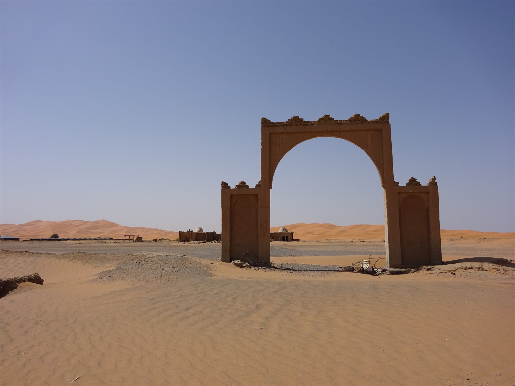 Adobe arch standing in Merzouga desert, dunes and houses under blue sky