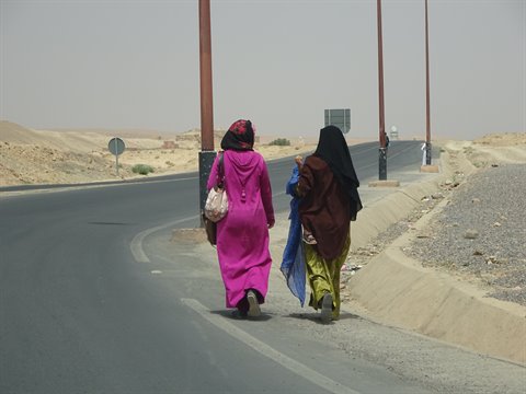 Women in djellabas walk along the asphalt road between sand and lampposts, Merzouga