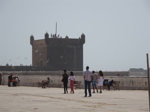 Tourists walk toward the Sqala du Port in Essaouira, seagulls in hazy sky