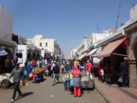 Open-air market among whitewashed arcades, crowds walking under colorful awnings, Essaouira