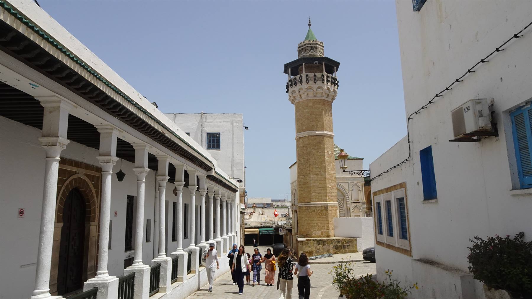 Turisti camminano lungo un vicolo con portici bianchi verso un minareto in pietra