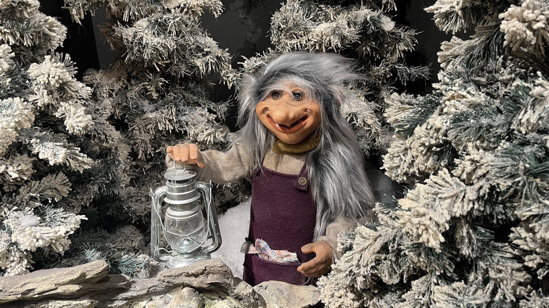 Statua di troll dai capelli grigi con lanterna tra abeti innevati