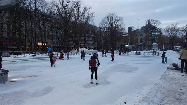 Skaters on a snowy ice rink in a city park under grey sky