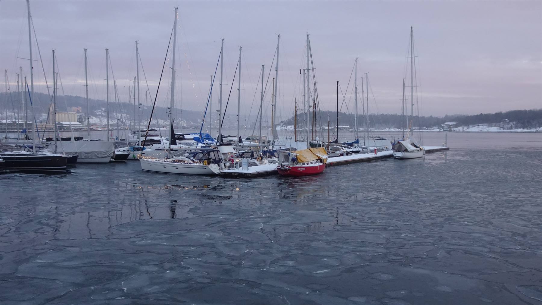 Barche a vela ormeggiate in un porto ghiacciato, neve sui ponti, luce invernale grigia