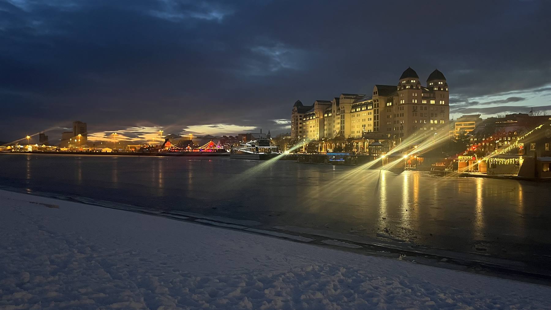 Neve sulla riva, edificio illuminato riflessa luci sull'acqua al crepuscolo