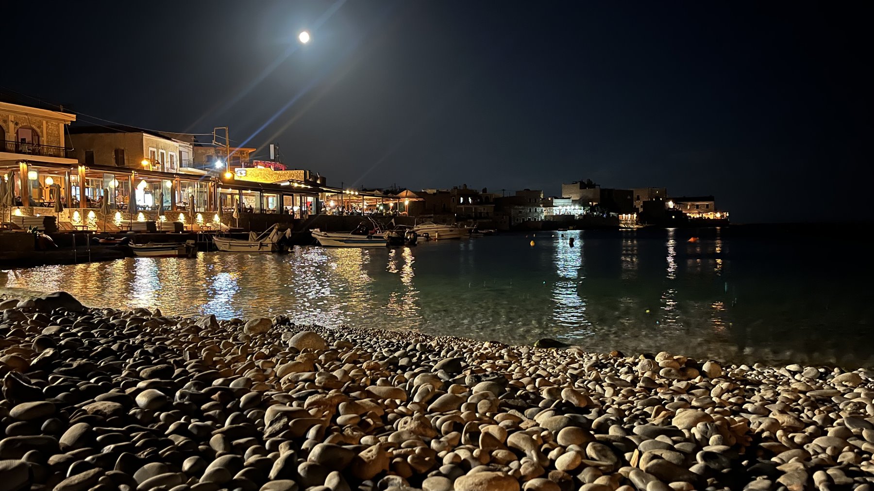 Una sera di luna piena, la spiaggia di sassi brilla sotto le luci del borgo.