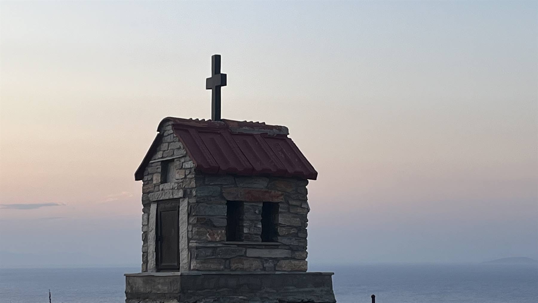 Piccola cappella in pietra con tetto rosso e croce, sul mare all'alba