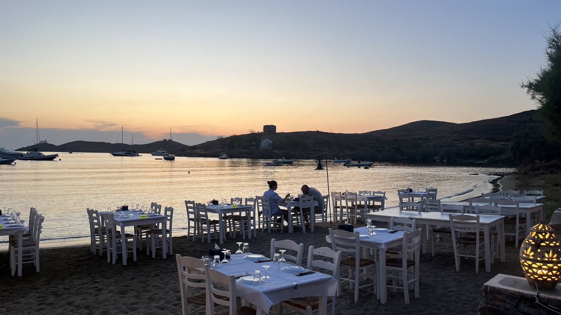 Tavole apparecchiate sulla spiaggia al crepuscolo, barche ancorate nella baia