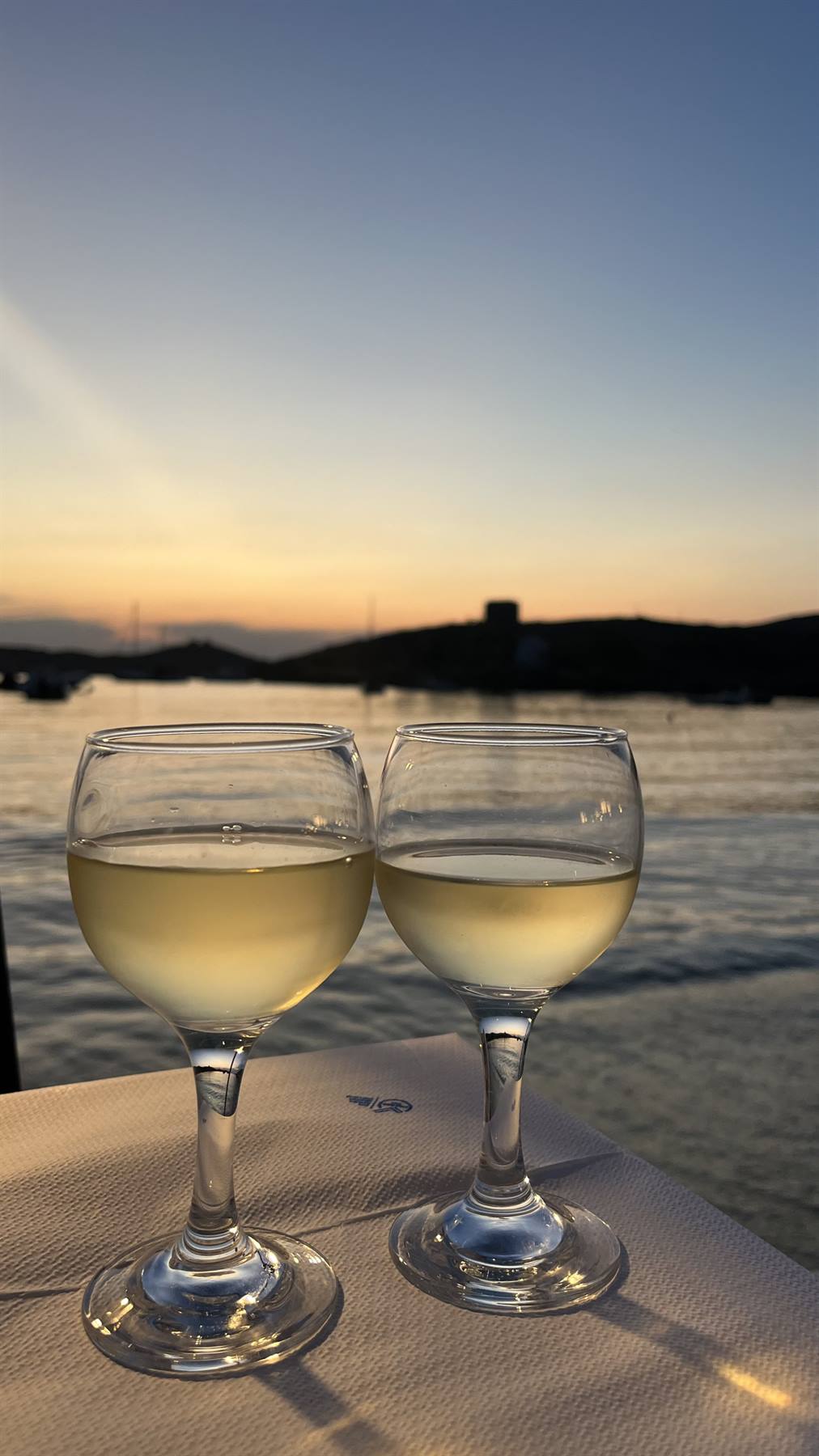 Due calici di vino bianco su tavolo, mare e colline al chiarore del crepuscolo
