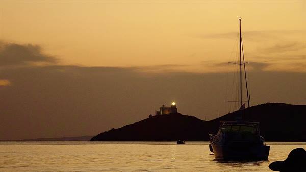Sailboat anchored offshore, lit lighthouse on dark hill against orange sky