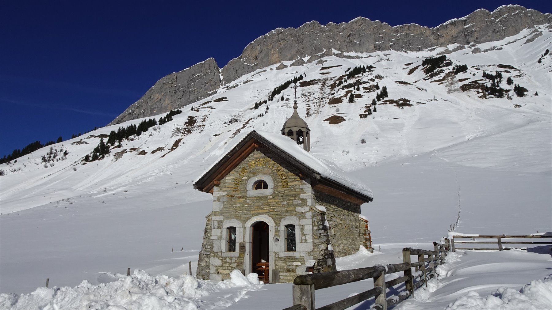Cappella in pietra innevata al Col des Aravis, sotto pareti rocciose e cielo blu