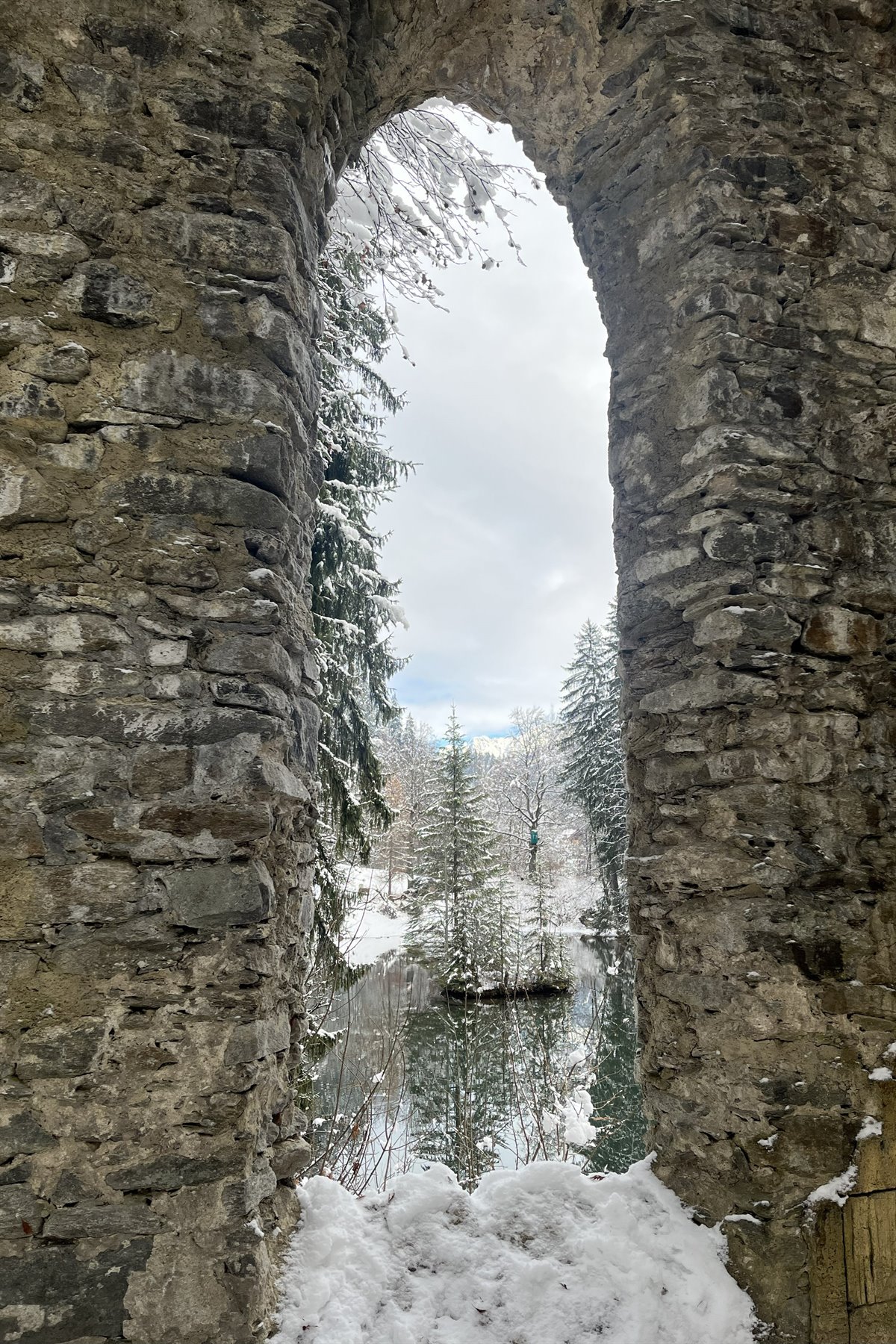 Arco di pietra antica innevata su alberi e laghetto gelido, Col des Aravis