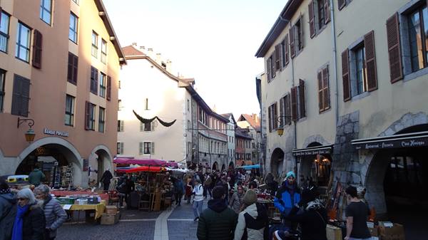 Market square in Annecy old town, shoppers browsing stalls under medieval arcades