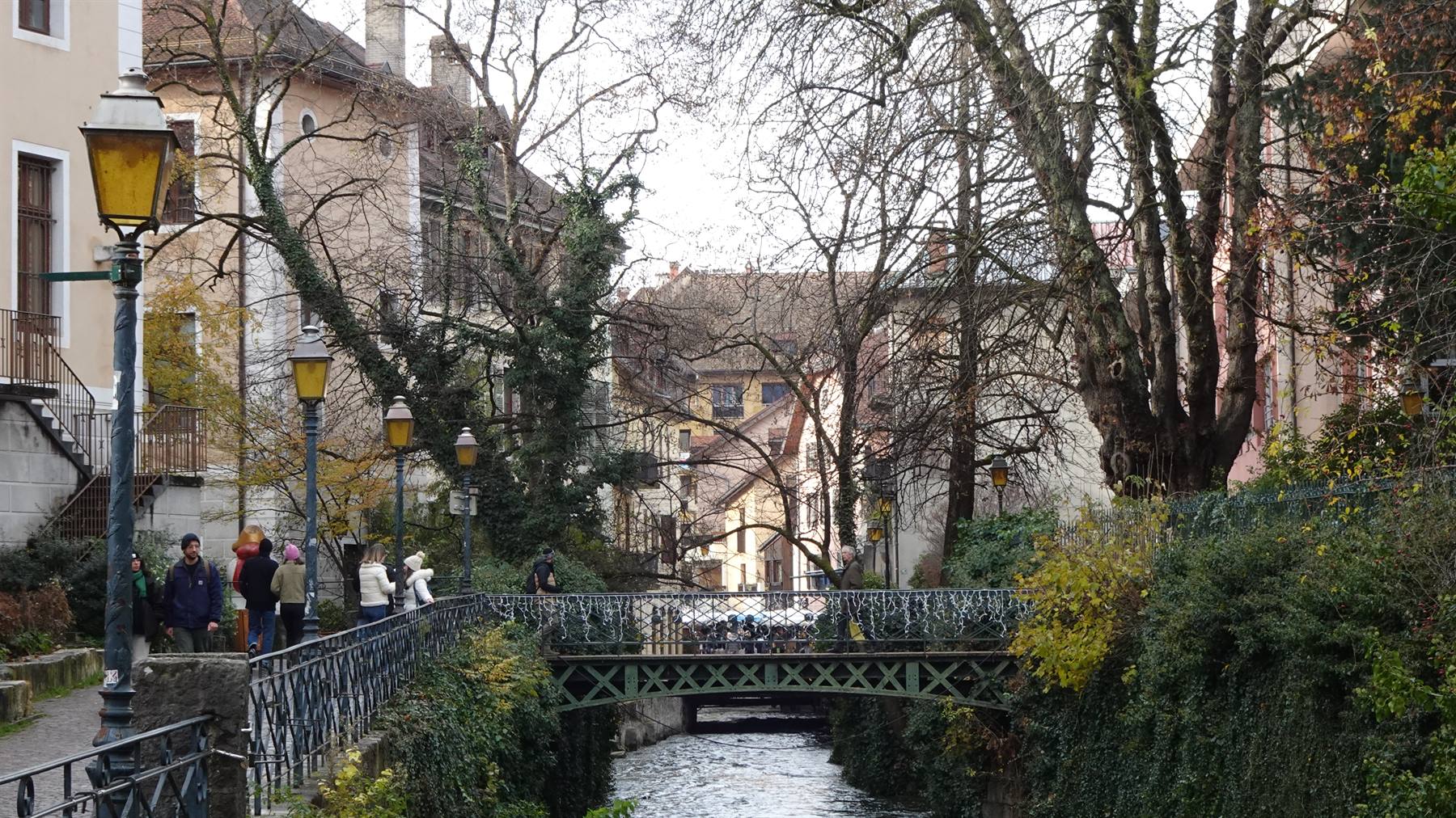 Turisti sul lungocanale ad Annecy, ponte di ferro in mezzo agli alberi spogli