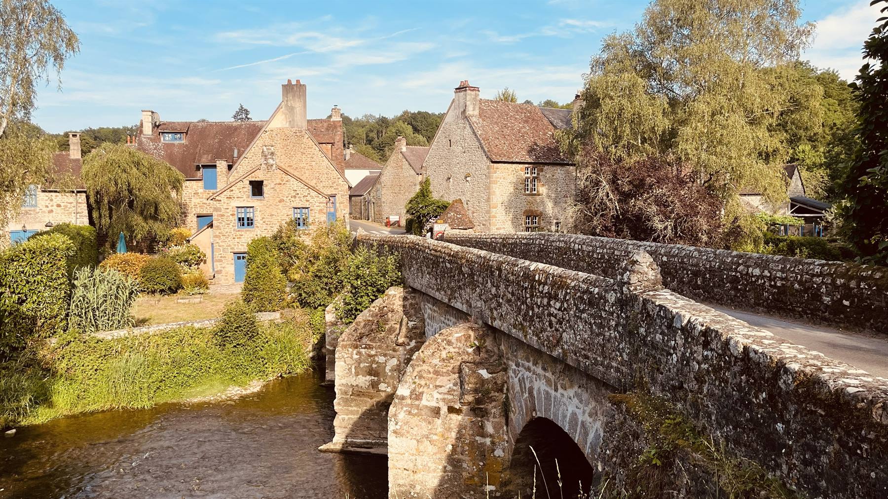 Ponte medievale in pietra sul fiume a Saint Céneri-le-Gérei, case e alberi soleggiati