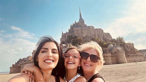 Three smiling women taking a selfie on the sand before Mont Saint Michel abbey