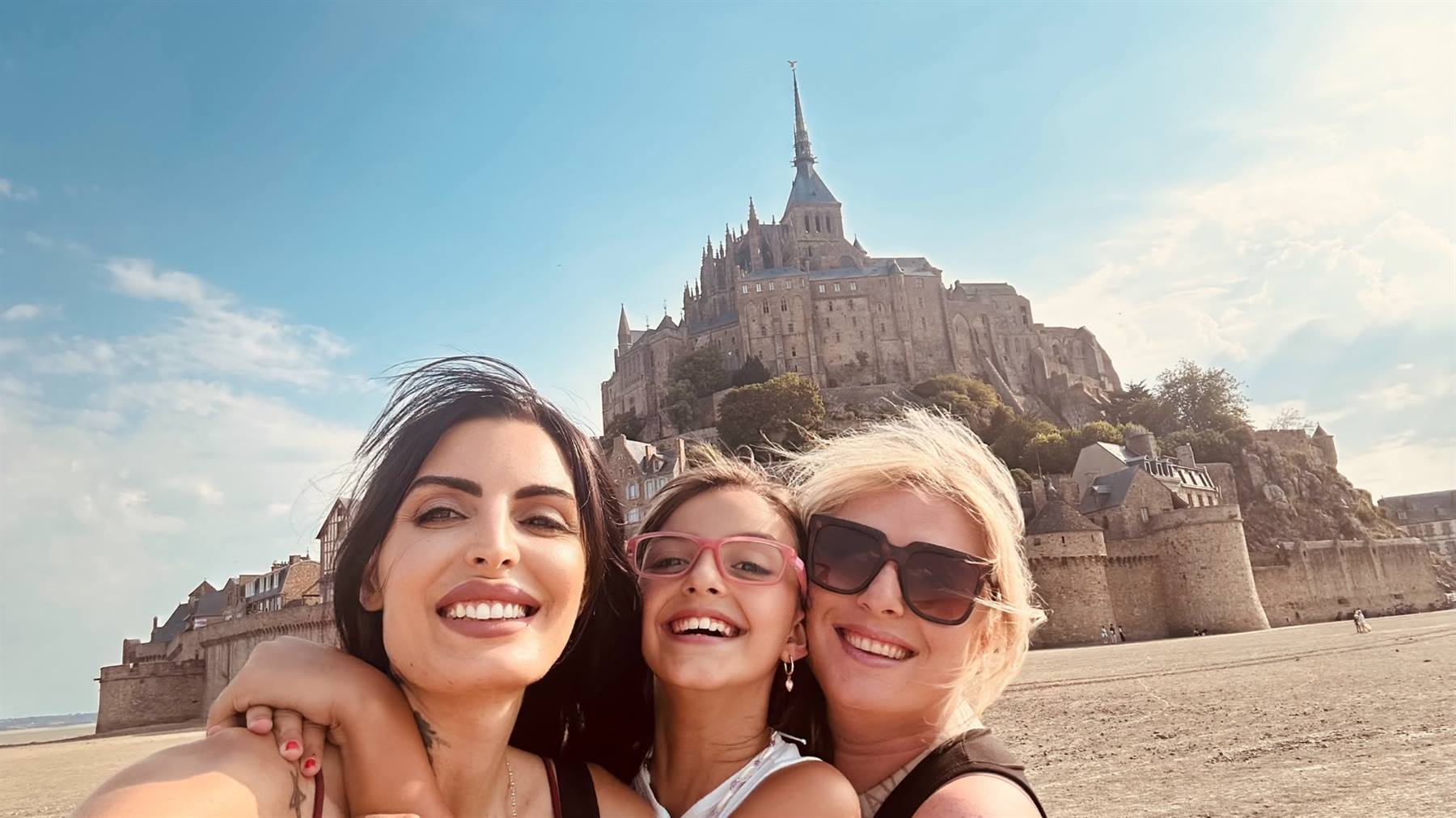 Selfie di tre donne sorridenti sulla sabbia davanti all'abbazia del Mont Saint Michel