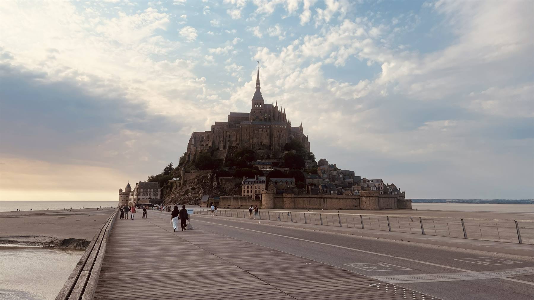 Visitatori camminano sul ponte verso l'abbazia di Mont Saint Michel tra sabbie e nuvole