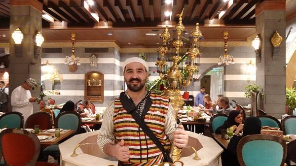Waiter in traditional striped vest smiles beside brass lamppost in restaurant
