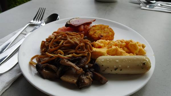 White plate with scrambled eggs, noodles, mushrooms and sausage, cutlery on table