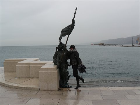 Tourist poses beside a bronze statue on the stone pier, grey sky