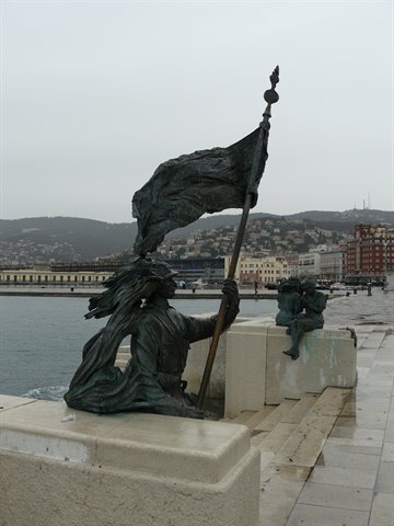 Bronze flag statue on Trieste pier, overcast sky, city and hills behind