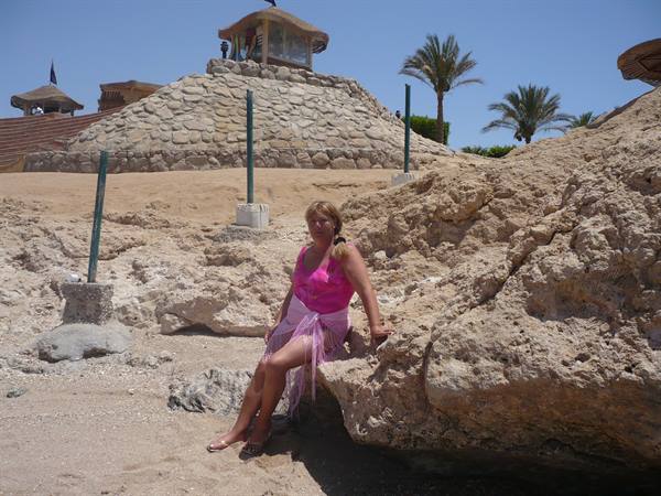 Woman in pink top sitting on rocky reef, palm trees and thatched kiosk behind