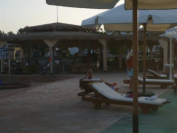 Woman lying on wooden sunbed by the pool, umbrellas and poolside bar behind