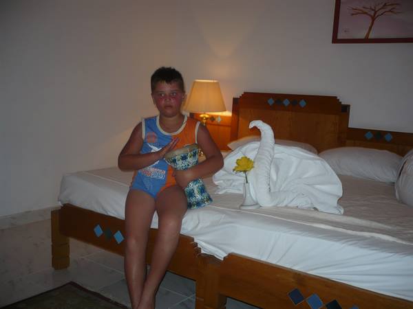 Boy in swimsuit sits on wooden bed next to towel swan and yellow flower