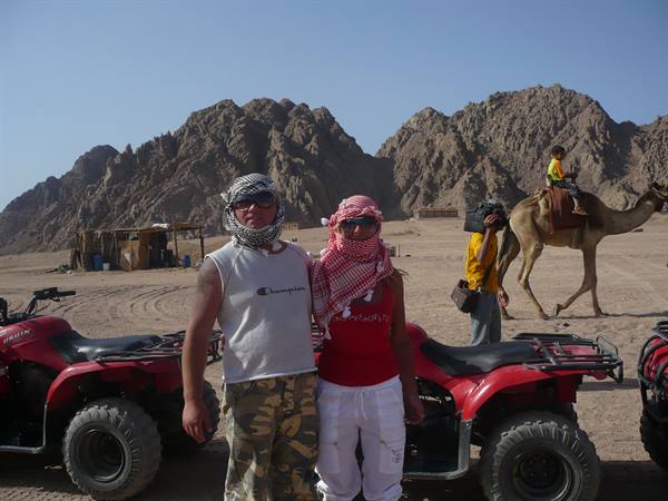 Tourists in keffiyehs beside red quad bikes in the desert, a camel passes rocky mountains
