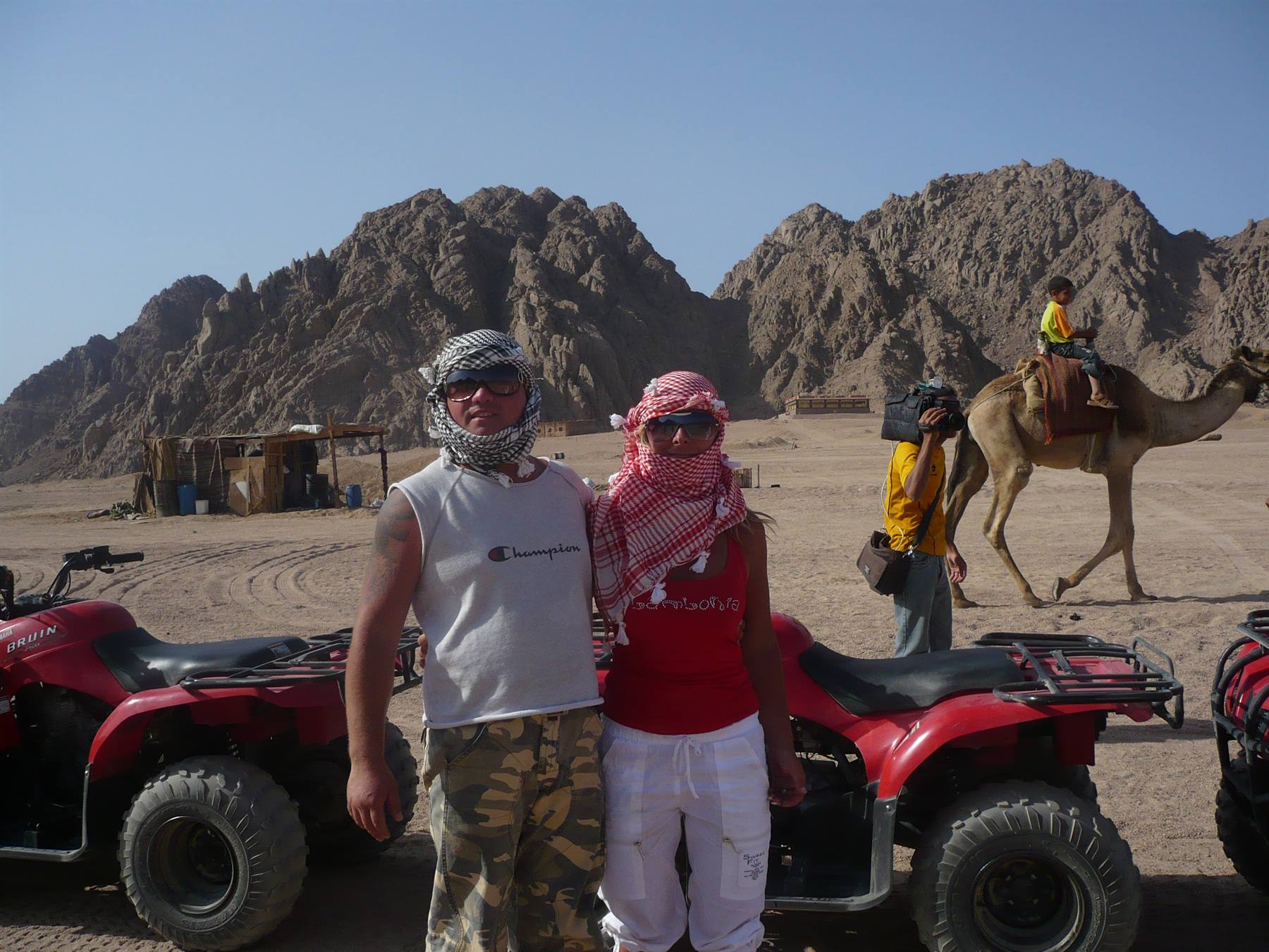 Tourists in keffiyehs beside red quad bikes in the desert, a camel passes rocky mountains