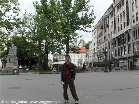 Tourist posing on a large paved square with monument, trees and historic buildings