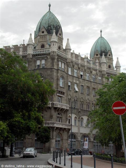 Historic stone building with green domes, trees and cars under grey sky