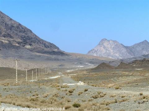 Dirt road crossing arid mountains, power line poles, blue sky, desert area