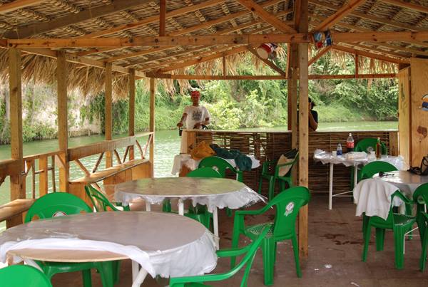 Tables with cloths and green chairs under thatched wooden shelter by the river