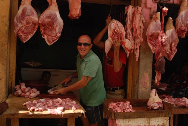Smiling butcher cuts meat under hanging carcasses at market stall