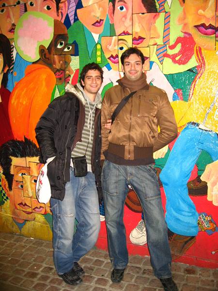 Two smiling young men in front of a colorful mural of painted faces