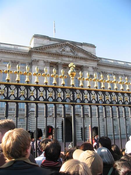 Tourists behind the golden railing at Buckingham Palace, royal guards under sunlight
