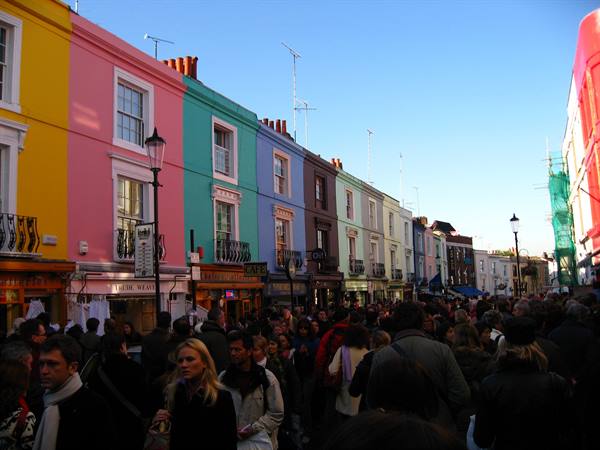 Crowd along Portobello Road past colorful terraced houses, lampposts and small shops
