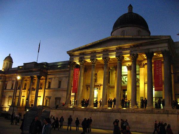 The National Gallery facade lit at night with columns and visitors at Trafalgar Square