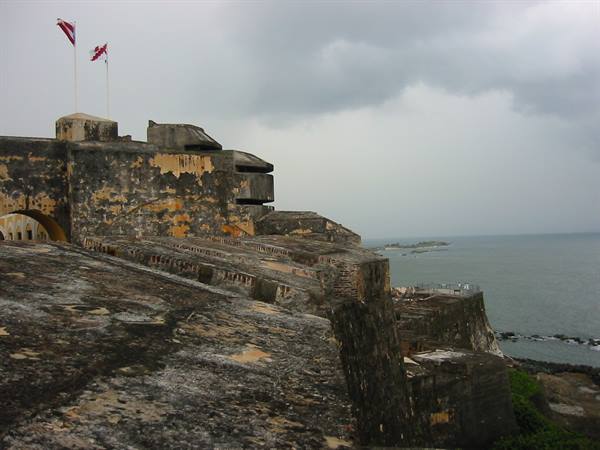 Stone walls of Castillo San Felipe del Morro with flags, ocean under cloudy sky