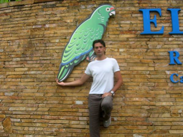 Man in white t-shirt poses next to a painted parrot on a brick wall