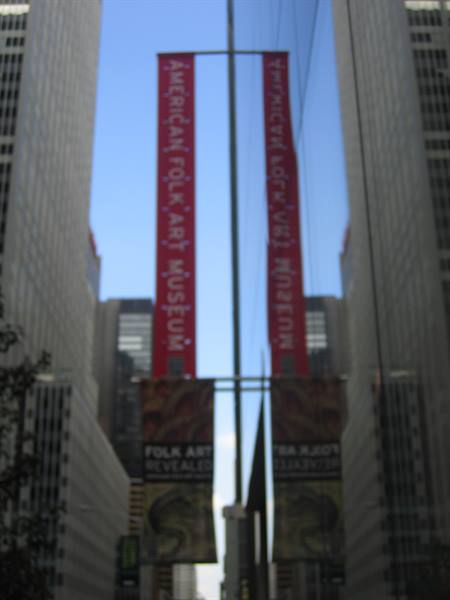 Red American Folk Art Museum banners among skyscrapers, reflected on glass