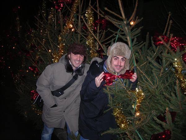 Two men in winter coats smile among decorated Christmas trees at night
