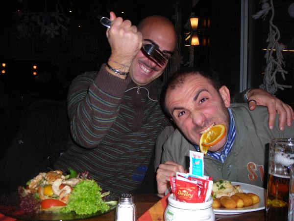 Two men laugh at a restaurant, one biting an orange slice